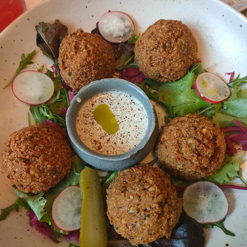 Cinco falafel à volta de uma taça de molho de tahini com sumac, com rabanete, pepino em picles e folhas
