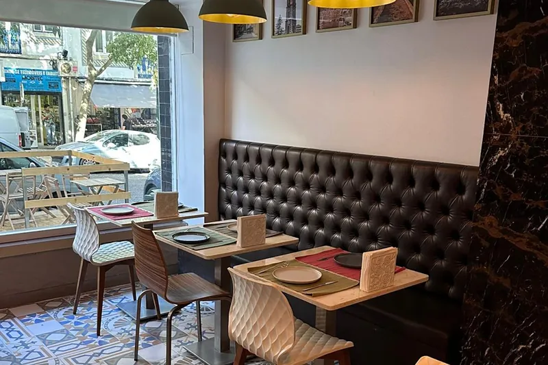 Mesas junto à montra, com um banco capitonado, toalhas individuais e o chão de mosaico; lá fora vê-se a esplanada